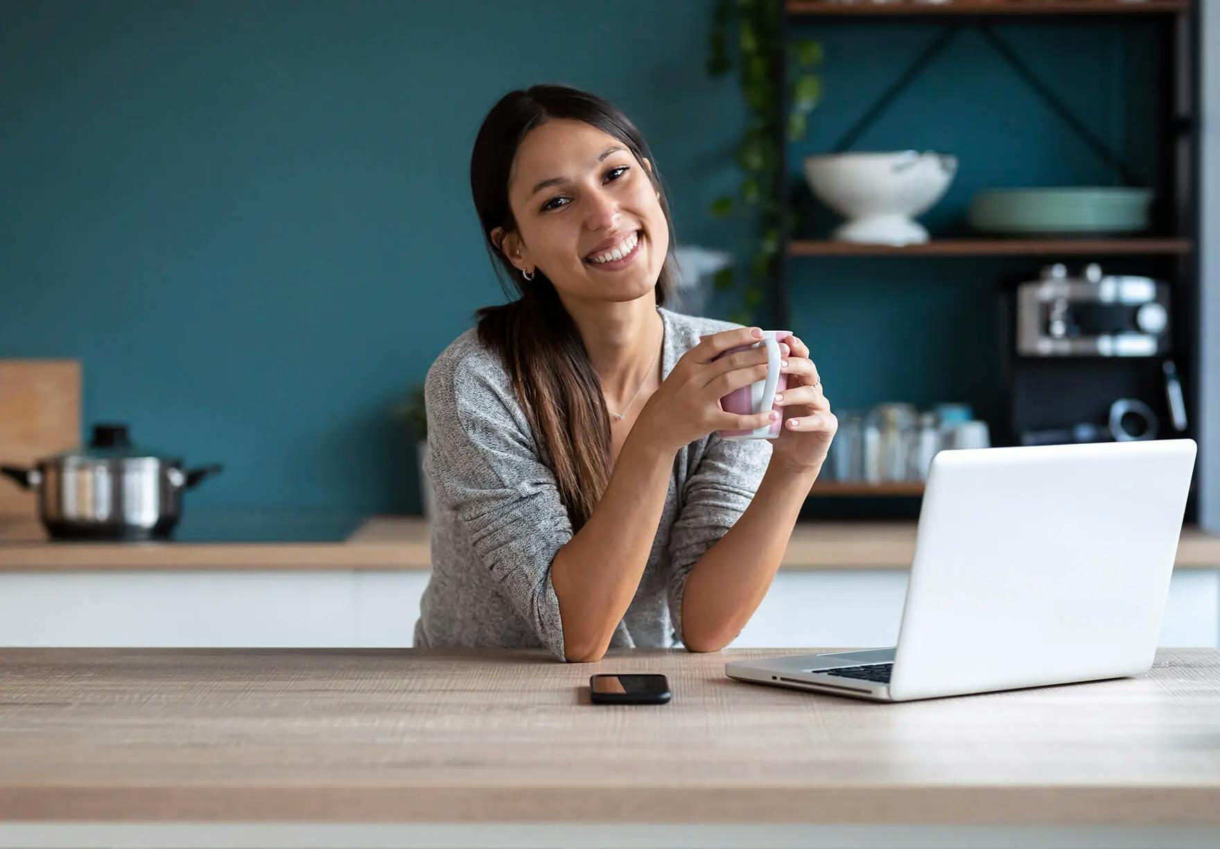 A woman sitting at a kitchen table with a laptop and a cup of coffee.