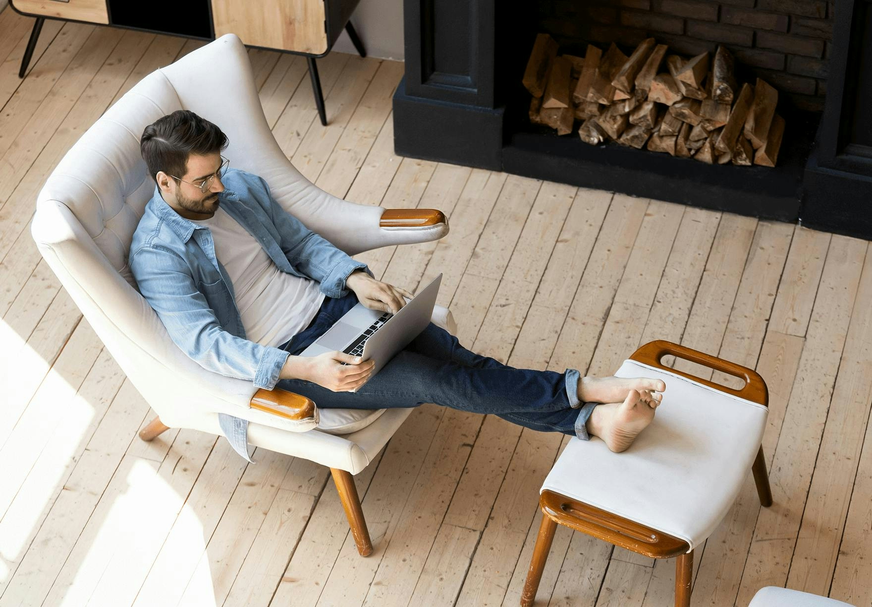a man sits comfortable in a chair by the fire while working on his laptop 